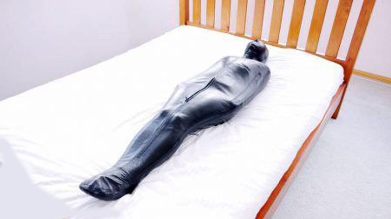 Full-length angled view of black leather hooded sleep sack laid on a bed, showing its tapered enclosed silhouette and front zipper detail.