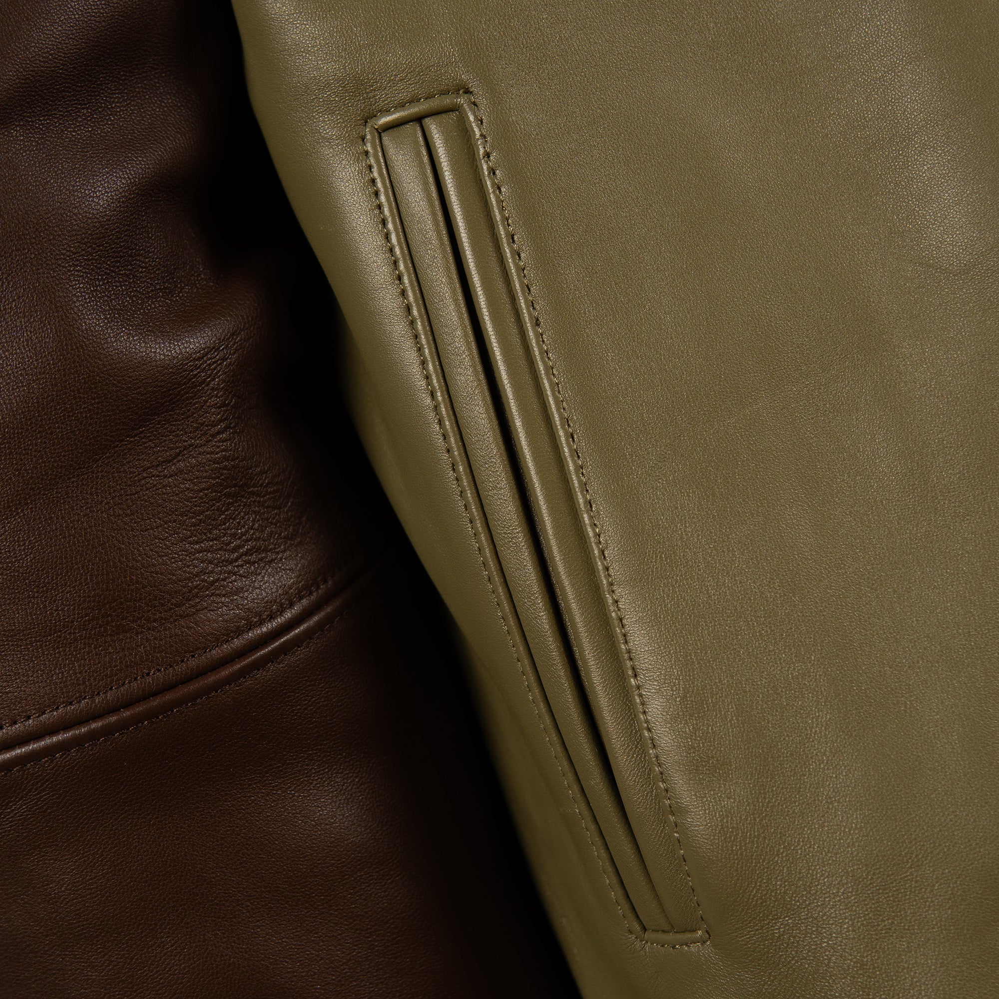 Close-up detail of a leather jacket showing a vertical welt pocket in olive green alongside a darker brown leather section.
