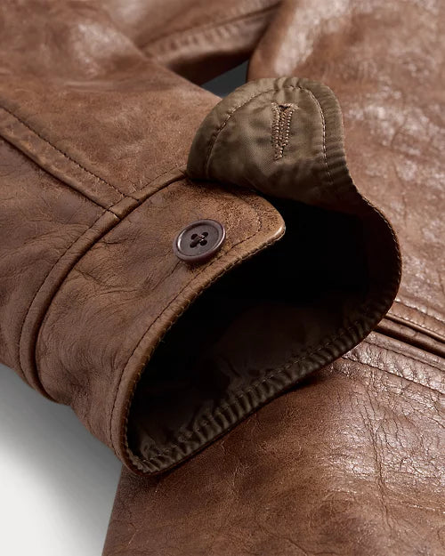 Close-up of a distressed brown leather jacket cuff showing its round button, stitched leather tab, contrasting inner facing and edge stitching.