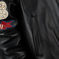 Women's black leather anniversary bomber jacket, front pocket detail, with red and blue crest artwork and decorative gold-toned accents.