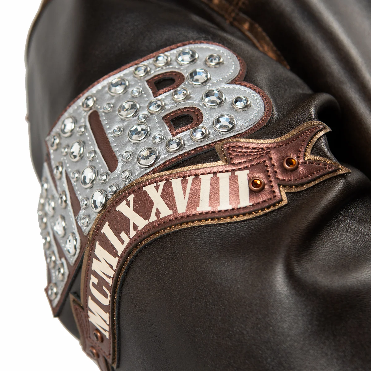 Women's distressed brown leather anniversary jacket, sleeve patch close-up, with cream lettering, layered crest artwork, and reflective embellishments.