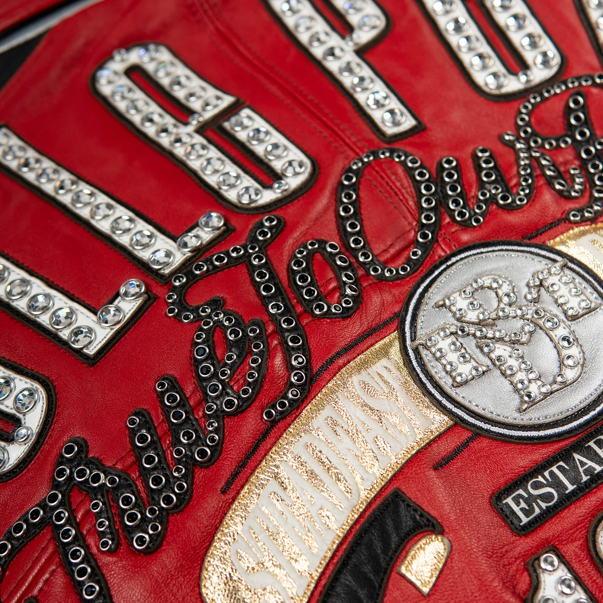 Women's red and black leather varsity jacket, back script and banner detail, with contrasting panels, embroidered motifs, and reflective embellishments.