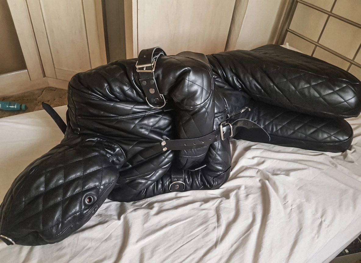 Black diamond quilted leather enclosed suit with matching hood, crossed covered sleeves and silver-tone buckle straps, displayed on a bed.