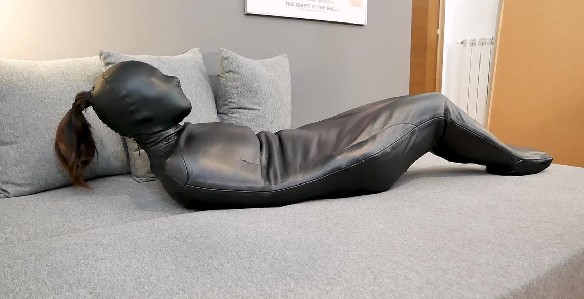 Side view of black genuine leather hooded sleep sack with contoured shoulder panels, enclosed body and foot section, shown on a gray sofa.