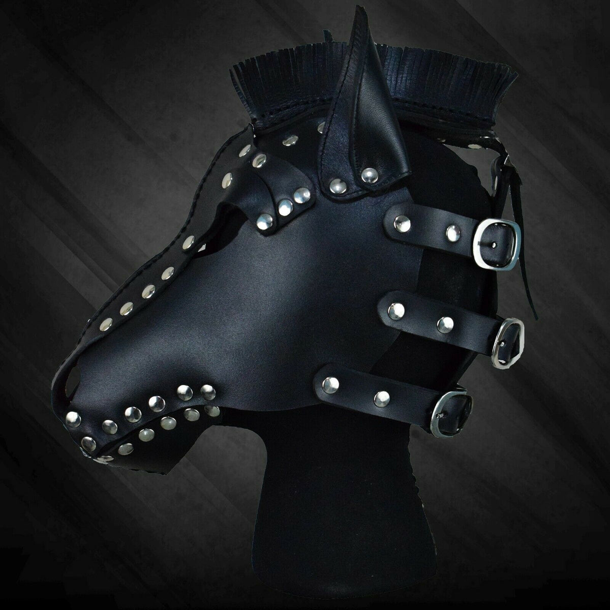 Side profile of black leather horse mask showing the long muzzle, pointed ear, fringe mane and three rear straps with silver-tone buckles.