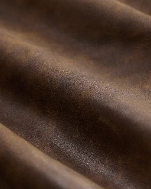 Detail of distressed brown genuine leather jacket material showing fine surface grain, warm tonal variation and soft natural folds.