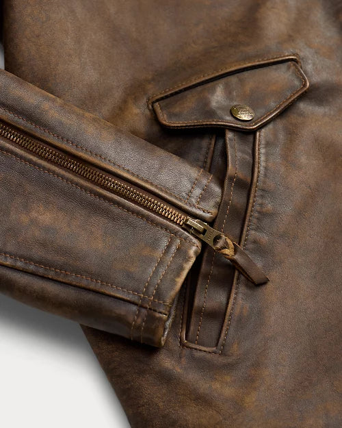 Close-up of distressed brown leather jacket showing a zip cuff with leather pull tab, stitched pocket opening and small snap flap.
