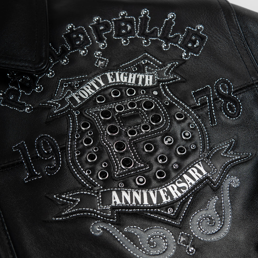 Chest crest detail on black leather jacket showing layered P applique, decorative eyelets, silver-tone stitching and ribbon-shaped patches.