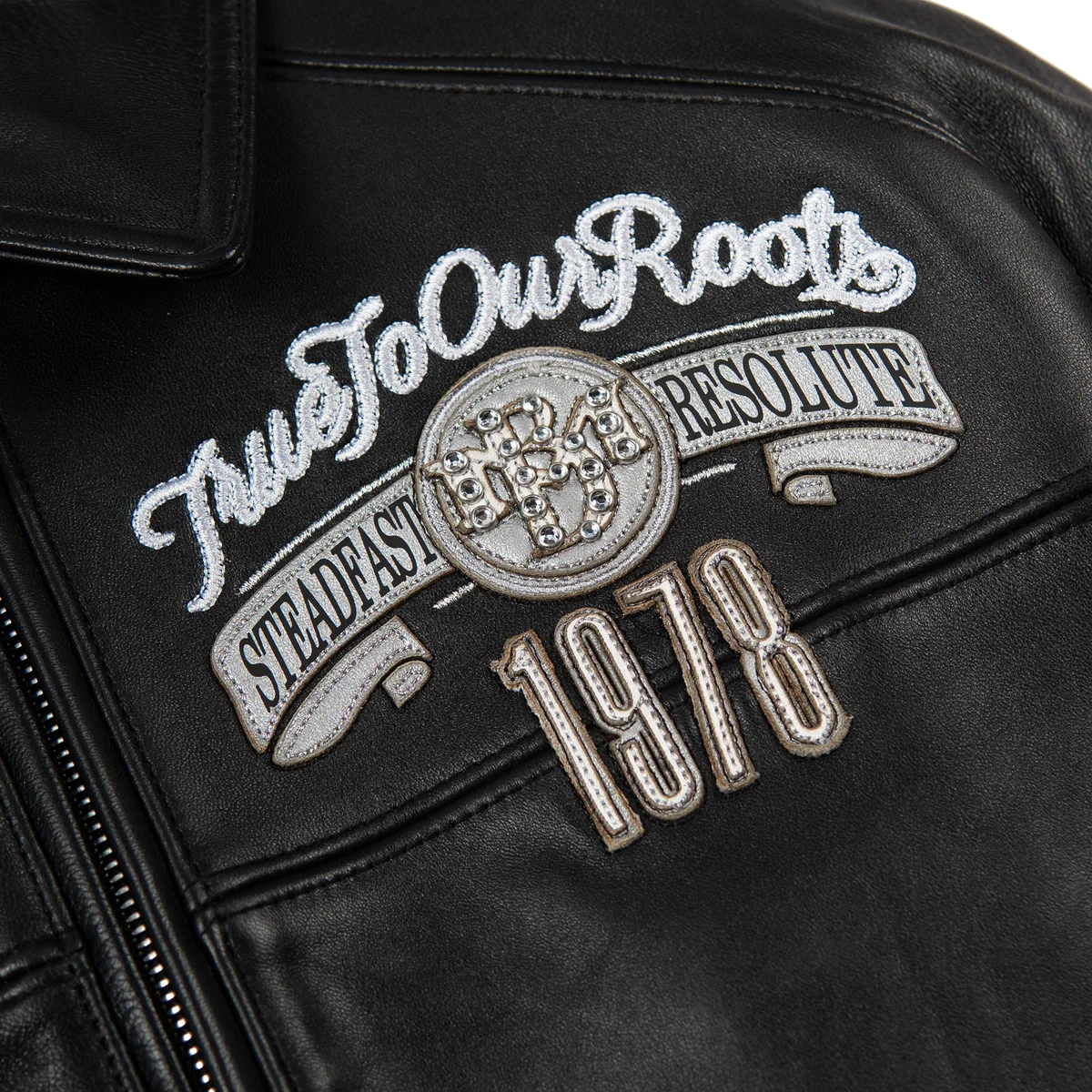 Chest detail of black leather jacket with white script embroidery, silver-tone medallion crest, ribbon applique and outlined 1978 numbers.