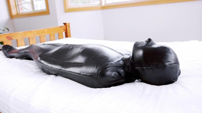 Profile view of black genuine leather sleep sack with a shaped hood, enclosed body and foot section, displayed horizontally on a white bed.