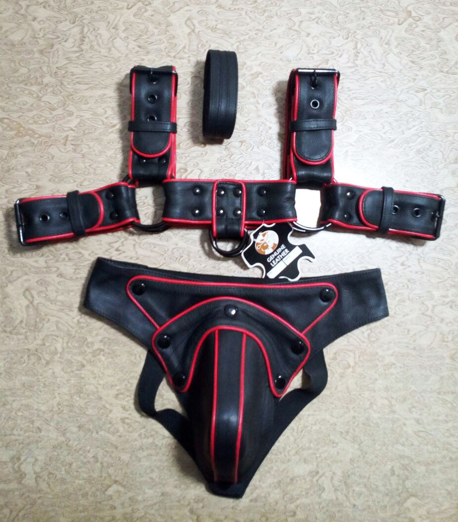 Black leather chest harness and matching jockstrap set laid flat, featuring red piping, buckle straps, metal rings and a shaped front cover.