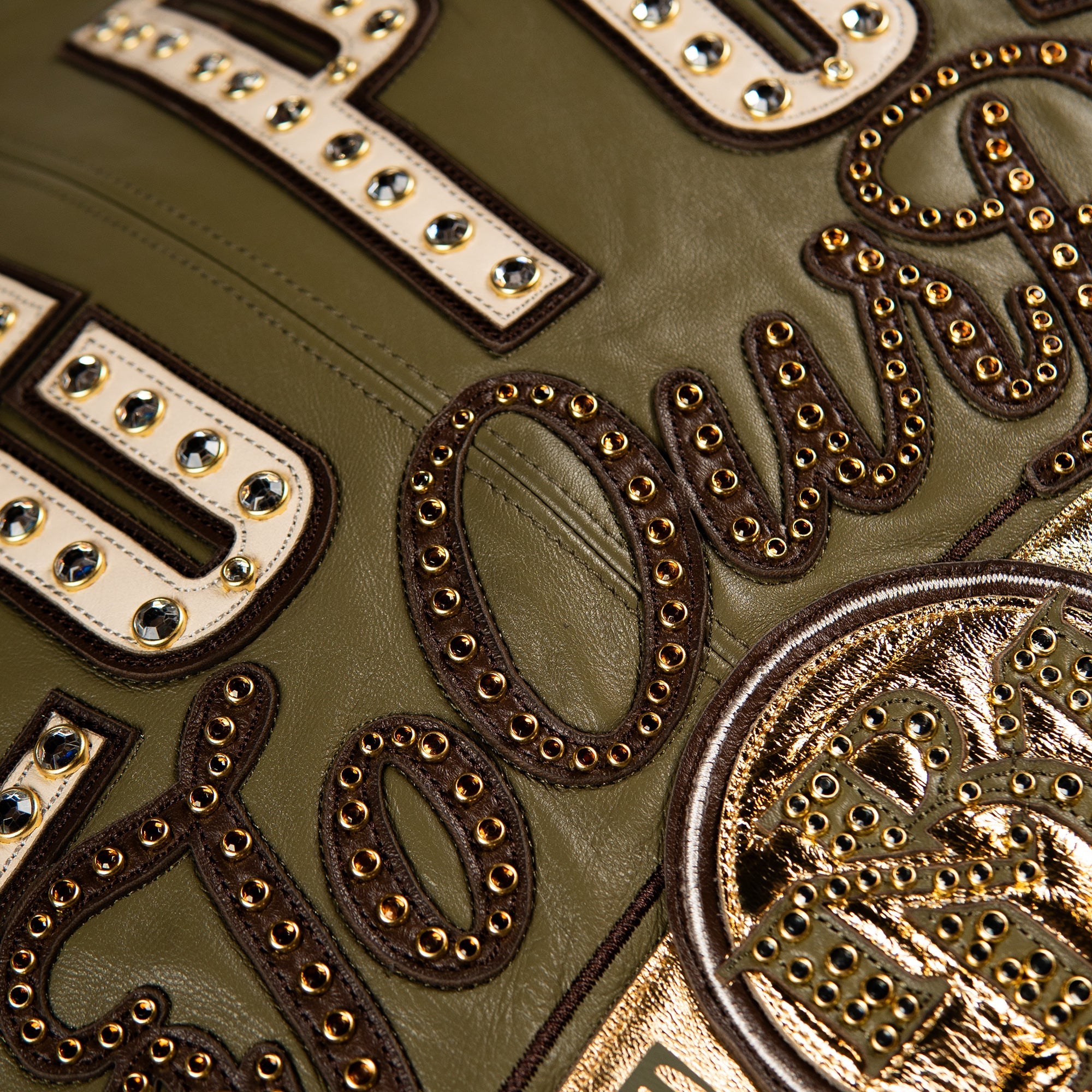 Close-up detail of a green leather jacket featuring large gold and white embroidered lettering embellished with silver studs and rhinestones.