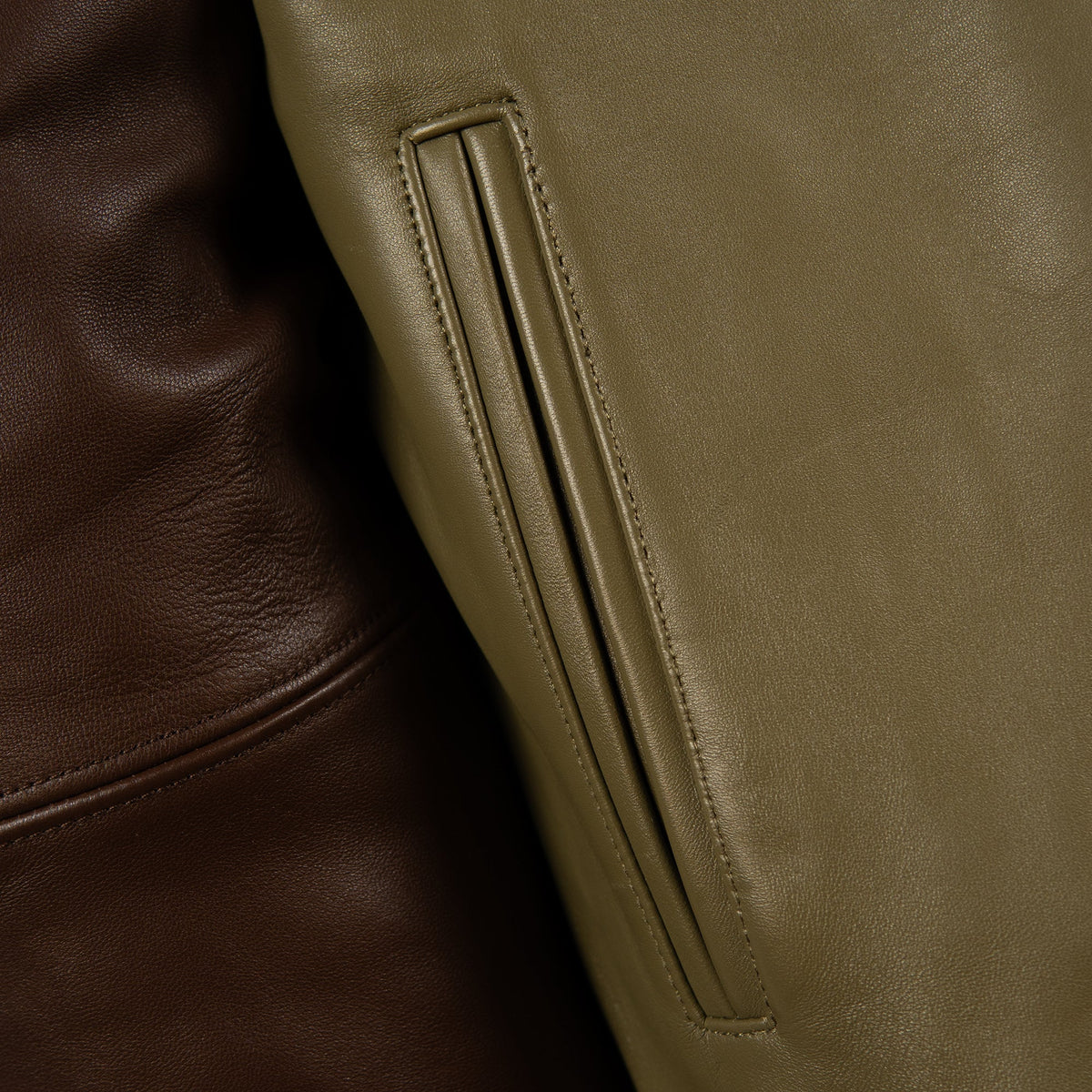Close-up detail of a leather jacket showing a vertical welt pocket in olive green alongside a darker brown leather section.