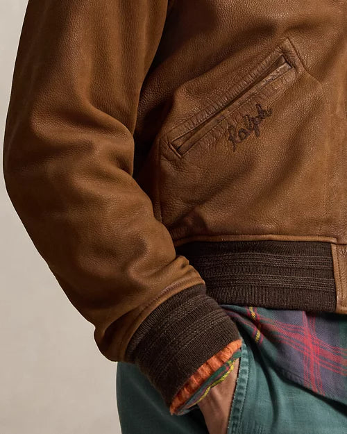 Close-up of a brown leather varsity jacket showing an angled welt pocket, embroidered branding, dark ribbed cuff and matching ribbed hem.