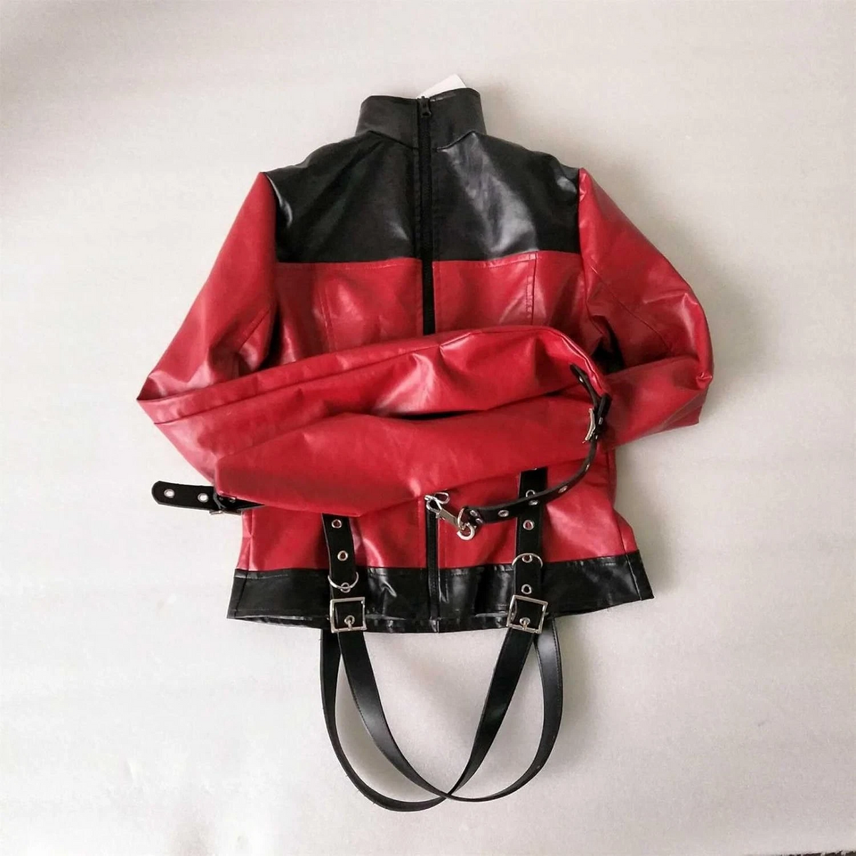 Red and black leather straitjacket laid flat with extended enclosed sleeves crossed across the body, rear zipper and black buckle straps.