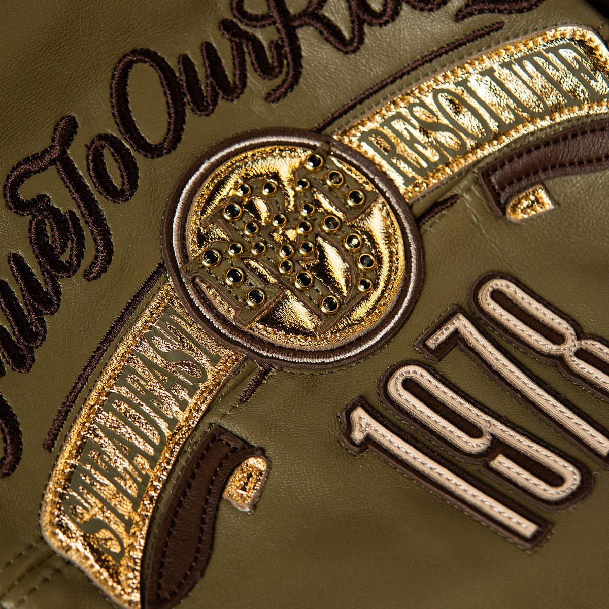 Close-up detail of an olive green leather patch with gold metallic embroidery, dark brown stitched lettering, and a circular emblem featuring a horse and rider.