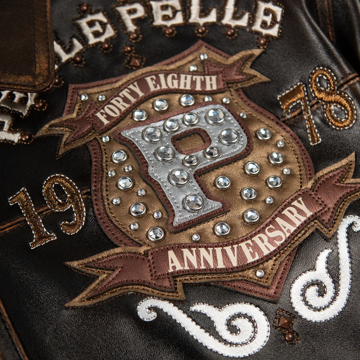 Women's distressed brown leather anniversary jacket, chest crest close-up, with cream lettering, layered crest artwork, and reflective embellishments.