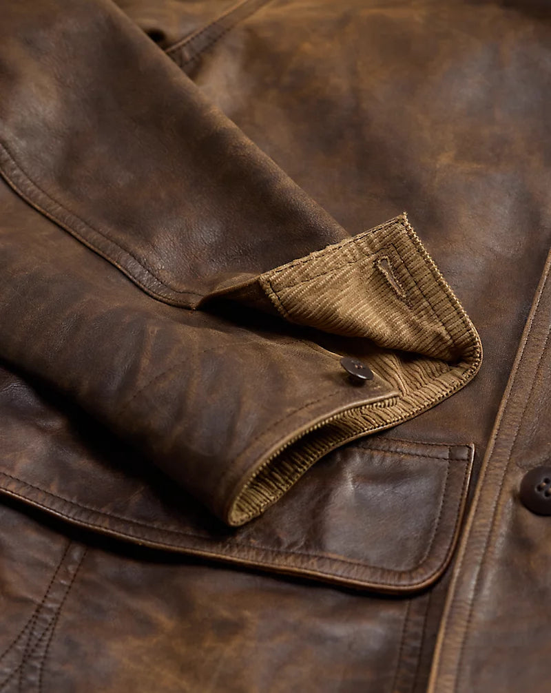 Close-up of brown leather jacket cuff showing a dark button, tan ribbed facing and stitched edges beside the front pocket flap.