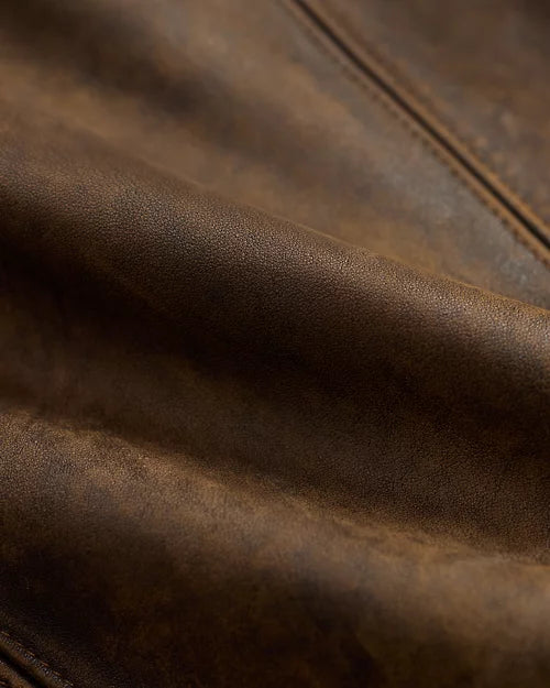 Detail of distressed brown genuine leather jacket material showing warm tonal variation, fine surface grain and a stitched panel edge.