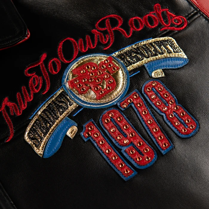 Close-up of True To Our Roots chest decoration with red script, gold-tone studs, blue edging and 1978 numerals on black leather.