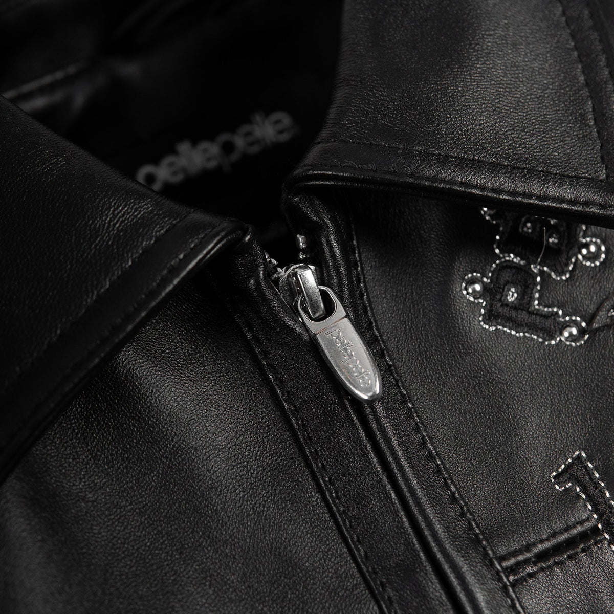 Close-up of black leather jacket collar and metal zipper pull showing leather grain, stitched edges and part of the chest embroidery.