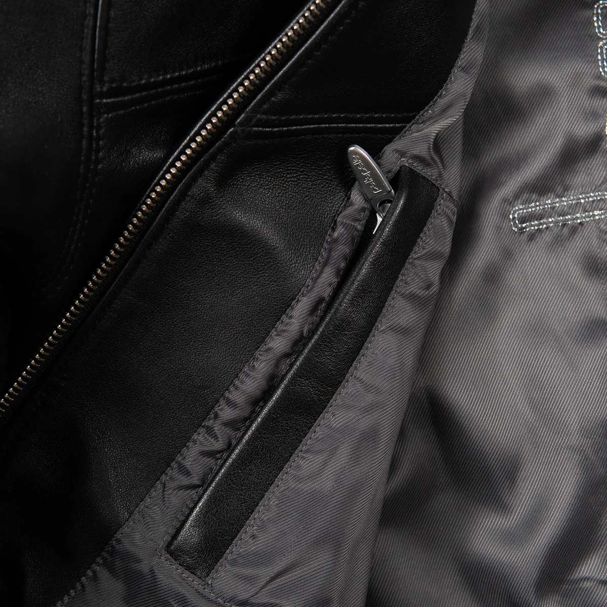 Interior detail of black leather jacket showing dark lining, leather-trimmed zip pocket, metal pull and central front zipper teeth.