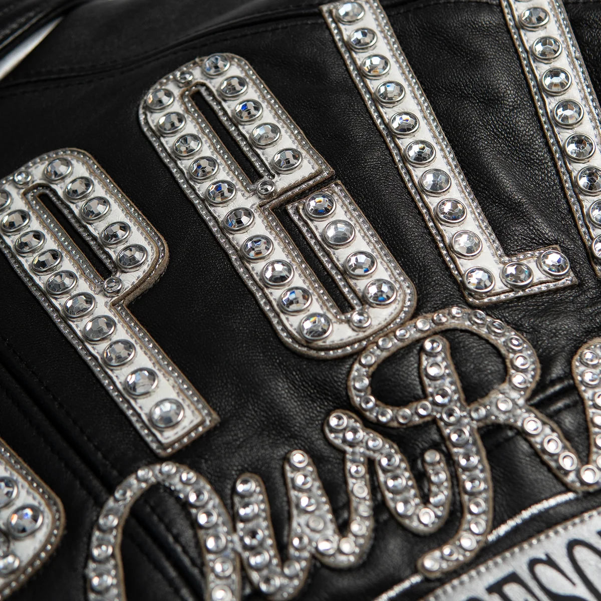 Close-up of silver-tone lettering on black leather jacket with rows of reflective decorative stones and stitched applique edges.