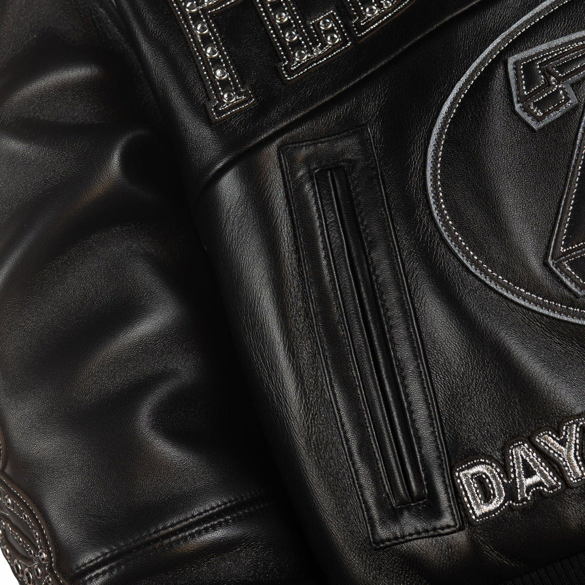 Detail of black leather jacket welt pocket beside large racing applique, decorative metal accents and silver-tone outlined lettering.