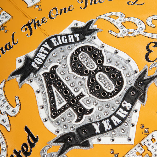 Close-up of large 48 back crest on yellow leather jacket with white and silver-tone panels, decorative eyelets and black ribbon shapes.