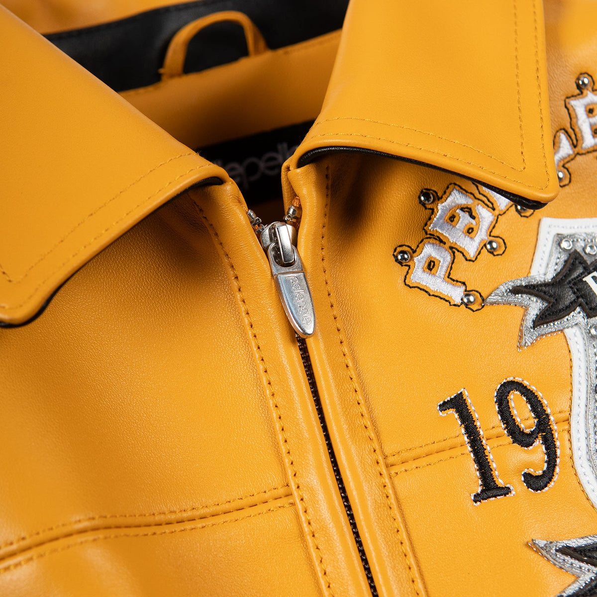 Collar detail of maize yellow genuine leather jacket showing pointed collar, stitched edges, metal zipper pull and chest embroidery.